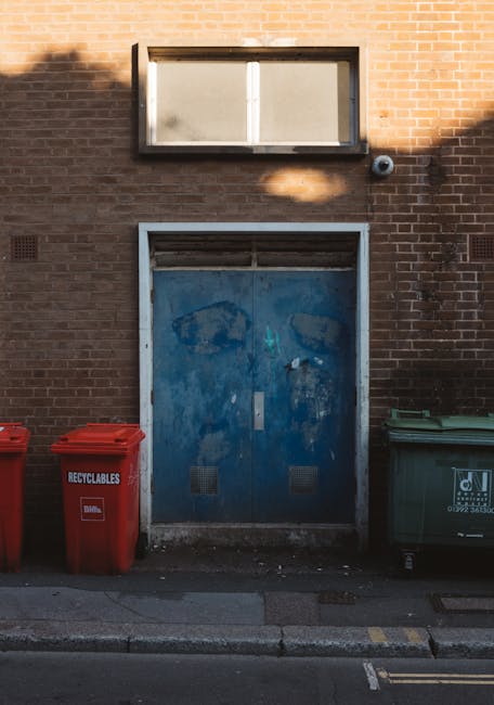 A narrow urban alleyway filled with discarded rubbish and debris, including a large black plastic bag and a wheeled refuse collection trolley in the foreground. The alley is bordered by tall, graffiti